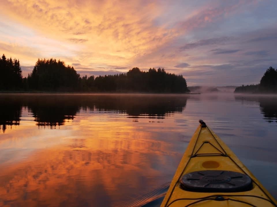 Joku meloo järvellä auringon laskiessa. Kuva on otettu kanootista ja siinä näkyy kanootin etuosa, järvi, pieni saari sekä laskeva aurinko.