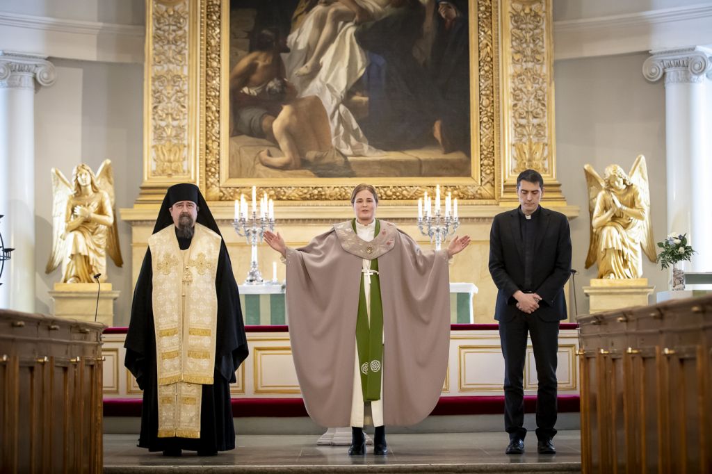 Piispa siunaa ihmisiä kirkon alttarilla. Piispan toisella puolella seisoo pappi ja toisella puolella ortodoksipiispa.