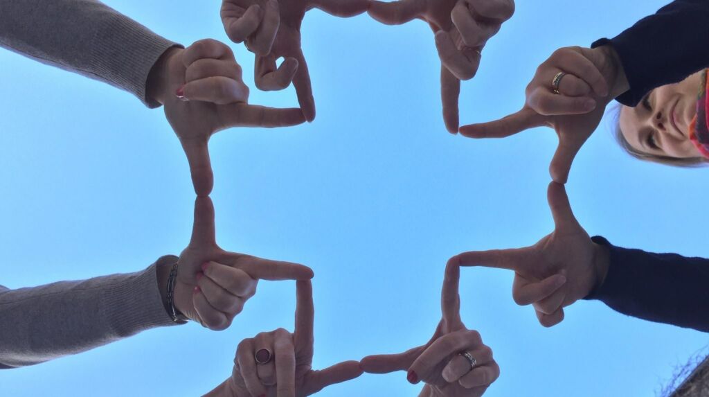 Neljä ihmistä muodostaa sormillaan ristin, taustalla sininen taivas