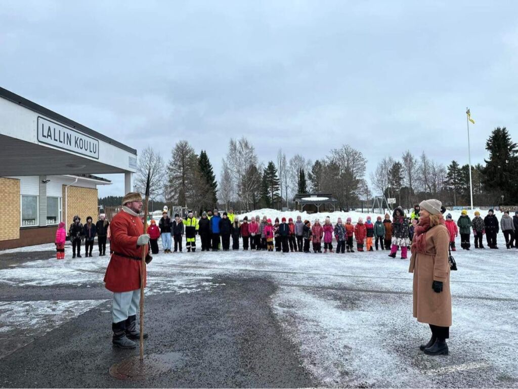 Kuvassa Lallin puvussa koulun rehtori, koululaisia ja piispa.